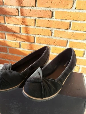 Graceland Svarta ballerinaskor med rosett 39 - Säljer ett par svarta ballerinaskor från Graceland med snygg rosettdetalj framtill. Skorna har rund tå och platt sula, tillverkade i mjuk mocka som ger en lyxig känsla. Perfekta att glida runt i när du vill ha en chill och stilren look.