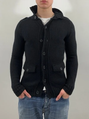 Sandro Stickad Cardigan Svart - Svart stickad cardigan från Sandro i slim fit. Modellen är 175 cm och väger 70 kg. Koftan passar S i slim fit. Inga defekter men tecken på användning som noppror och dammansamling. Nypris ca 3000 kr.