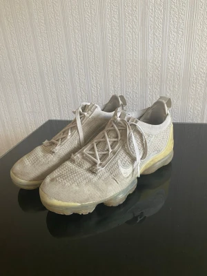 Beige Nike Vapormax sneakers - Nike Vapormax sneakers i beige med stickad ovandel och transparent sula. Skorna har snörning och en rund tå, samt en lätt och flexibel design. Swoosh-loggan syns på sidan och hälen har en förstärkt detalj. Perfekt för dig som gillar sportig stil.