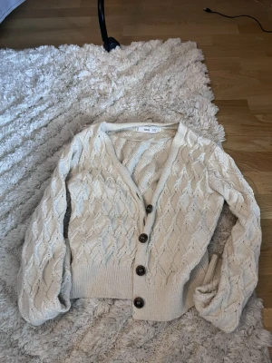 Beige stickad kofta från Mango - Säljer en beige stickad kofta från Mango med snyggt kabelstickat mönster och stora svarta knappar framtill. Koftan har v-ringad hals och långa ärmar med ribbade muddar. Perfekt för lager-på-lager och chill dagar.