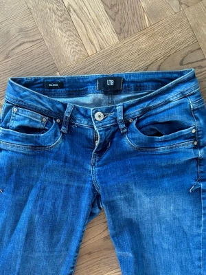 Blå bootcut jeans från LTB W26 L32 - Snygga blå jeans från LTB i bootcut-modell med klassisk femficksdesign och slitningar för en cool look. Jeansen har låg midja och är tillverkade i jeansmaterial med kontrastsömmar och detaljerade bakfickor.