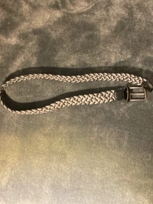 Gråsvart flätad bälte med skinn - Säljer ett snyggt flätat bälte i grått och svart med detaljer i svart skinn och metallspänne. Bältet har ett coolt mönster och passar perfekt till jeans eller chinos. Längden är 80 cm och materialet är en mix av syntet och skinn.