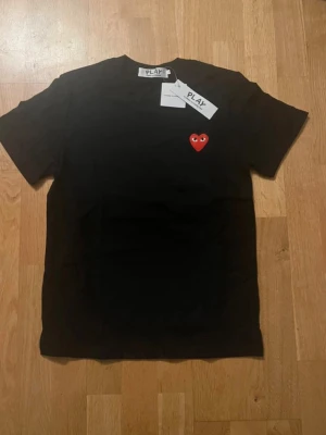 Svart t-shirt Comme des Garçons Play - Svart t-shirt från Comme des Garçons Play med klassiskt rött hjärta med ögon broderat på bröstet. T-shirten är kortärmad och har en rund hals. Materialet är mjuk bomull och passformen är normal. Perfekt för dig som gillar stilrena och ikoniska plagg.