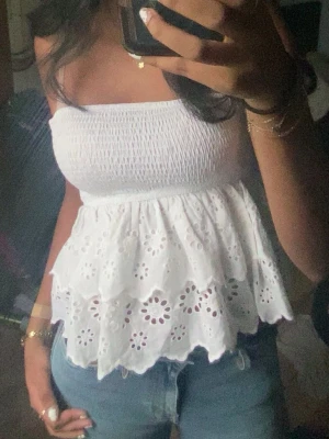 Vit axelbandslös broderad topp - Säljer en söt vit axelbandslös topp med smock upptill och dubbla volanger med broderade blommönster. Toppen har en romantisk känsla och är perfekt till sommaren. Materialet känns lätt och luftigt, och den har en bandeauform som sitter snyggt.