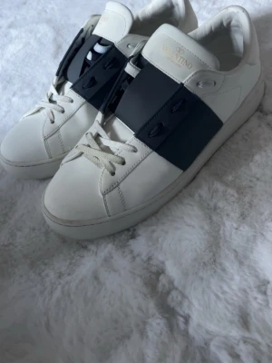 Valentino Garavani Open sneakers vit/blå - Säljer ett par Valentino Garavani Open sneakers i vitt skinn med bred marinblå detalj över vristen. Klassisk rund tå, vita snören och diskret logga på plösen. Snygg och stilren design som passar till allt. Kommer med originalkartong, extra skosnören och dustbag.
