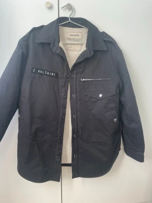 Svart overshirt från Zadig & Voltaire - Svart overshirt från Zadig & Voltaire med vit teddyfoder på insidan. Jackan har knappar framtill, en bröstficka med dragkedja och patch med Z. Voltaire. Perfekt för lager-på-lager och har en cool vintagekänsla. Ganska rymlig. 