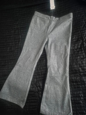 Glittriga bootcut byxor i silver Barn - Säljer ett par glittriga bootcut-byxor i silver med snyggt skimmer. Byxorna är tillverkade i en ekologisk bomullsblandning och har en bekväm midja. Perfekta för dig som vill sticka ut med en cool look och gillar metallic-effekt.