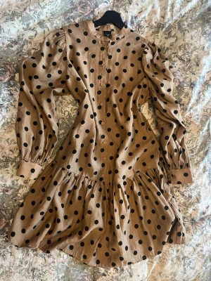 Prickig kortklänning från ONLY Studio - Snygg beige kortklänning med svarta prickar från ONLY Studio. Klänningen har långärmade puffärmar, rund hals och knappar hela vägen framtill. Tillverkad i ett mjukt och lätt material som ger en skön känsla. Perfekt för dig som gillar trendiga mönster och voluminösa detaljer. Klänningen har alldrig använts. 