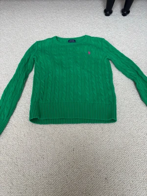 Grön stickad tröja från Polo Ralph Lauren - Snygg grön kabelstickad tröja från Polo Ralph Lauren med rund hals och lång ärm. Tröjan har ett rosa broderat Polo-logo på bröstet och breda ribbade muddar vid ärmslut och nederkant. Perfekt för dig som gillar klassisk stil med färg.