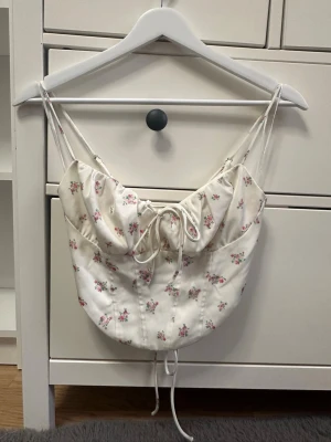 Vit korsett-topp med blommigt mönster - Superfin korsett-topp i vit färg med små rosa och gröna blommor. Toppen har smala axelband, snörning både fram och bak samt en croppad passform. Materialet känns mjukt och har en lätt glans. Perfekt för dig som gillar romantiska detaljer och vill sticka ut.