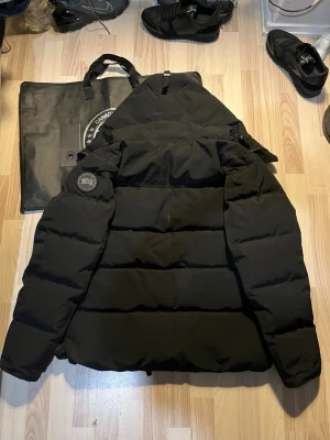Svart pufferjacka från Canada Goose - Svart pufferjacka från Canada Goose med huva och snyggt märke på ärmen. Jackan har dragkedja framtill och är fodrad för extra värme. Perfekt för kalla dagar och har en clean, stilren look som funkar till streetwear. Modellen är Fusion L/G.