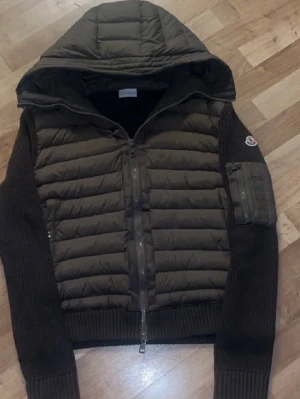 Moncler cardigan - Säljer en mörkbrun pufferjacka från Moncler med stickade ärmar och huva. Jackan har dragkedja framtill, ribbade muddar och en ficka med dragkedja på ärmen. Perfekt för dig som vill ha en snygg och varm jacka med coola detaljer.