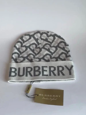 BB mössa - Snygg mössa från BB i ljusgrått och vitt med ett coolt hjärtmönster och stor Burberry-logga framtill. Stickad i mjukt material som känns skönt mot huvudet. Perfekt för dig som vill ha en trendig accessoar med lyxig känsla.