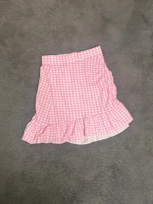 Rutig rosa volangkjol Gina Tricot - Supersöt kort kjol från Gina Tricot i rosa och vitt rutigt mönster. Kjolen har volang längst ner och är gjord i mjuk bomull, perfekt för dig som gillar en lekfull och trendig stil. Passar dig som vill ha en feminin och fräsch look.