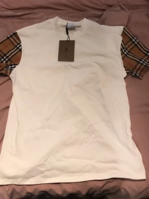 Vit Burberry t-shirt med rutiga ärmar - Säljer en clean vit t-shirt från Burberry med klassiska rutiga ärmar i beige, svart och rött. T-shirten har rund hals och är kortärmad, tillverkad i mjuk bomull. Perfekt för dig som gillar stilrena plagg med en twist från ett ikoniskt märke.