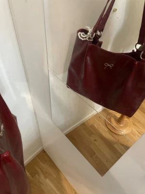 Vinröd axelväska med pärldetalj - Säljer en vinröd axelväska i glansigt skinnimitation med dubbla axelband och silverfärgade ringar. Väskan har en söt metallrosett framtill och en pärlrosett som dekoration. Perfekt storlek för allt du behöver ha med dig och ger en chic vibe till din outfit.