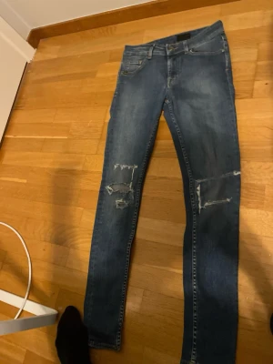  jeans med slitningar - Säljer ett par blå skinny jeans med snygga slitningar på båda benen. Klassisk femficksmodell med gul söm på bakfickorna. Jeansen har en smal passform och är tillverkade i jeansmaterial med lite stretch för extra komfort.