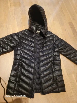 Svart dunjacka från McKinley - Säljer en svart dunjacka från McKinley med huva och snyggt quiltat mönster. Jackan har dragkedja framtill och reflekterande detaljer nedtill på ryggen. Perfekt för kalla dagar och riktigt skön att ha på sig.