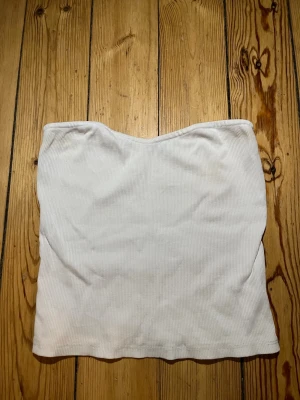Vit ribbad tubtopp från Gina Tricot - Säljer en vit ribbad tubtopp från Gina Tricot. Toppen är axelbandslös och har en enkel, stilren design med lätt böjd överkant. Perfekt att matcha med jeans eller kjol för en fräsch look.
