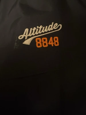 Svart vindjacka från 8848 Altitude - Snygg svart vindjacka från 8848 Altitude med vita detaljer och gråa ärmar. Jackan har dragkedja framtill, två fickor med dragkedja och broderad logga på bröstet. Sportig look med randiga muddar och meshfoder på insidan.