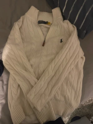 Knitsweater Polo Ralph Lauren - Säljer en vit kabelstickad tröja från Polo Ralph Lauren i storlek M. Tröjan har half zip med brunt läderdrag, ribbade muddar och klassisk broderad logga på bröstet. Perfekt för lager-på-lager och chill stil.