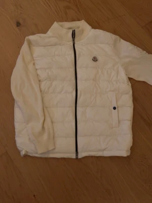 Moncler Vit Cardigan  - Säljer en snygg vit cardigan från Moncler med stickade ärmar och baksida. broderad Moncler-logga på bröstet samt ett mönster på ärmen. Jättefin och använd endast 2-3 gånger, då den passar lite kort på mig nu o kommer ej passa mig till sommaren. Den passar typ M / L.