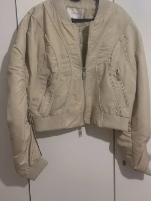 Beige bomberjacka från Gestuz - Snygg beige bomberjacka från Gestuz med croppad passform och ribbade muddar. Jackan har två framfickor med dragkedja och coola sömdetaljer som ger en modern vibe. Perfekt att slänga över en hoodie eller t-shirt för en chill look.