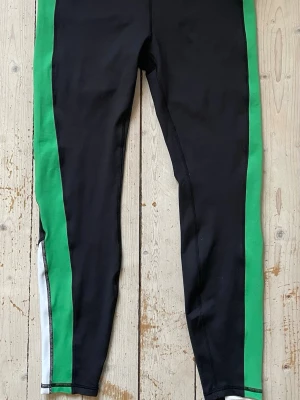 Svarta träningstights från Stronger stl. L - Snygga svarta träningstights från Stronger med gröna paneler längs sidorna och vita detaljer vid anklarna. Tillverkade i stretchigt material som passar perfekt för träning och sportiga aktiviteter. Oanvända/nyskick. 