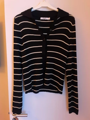 Svartvit randig kofta från NA-KD - Säljer en svart kofta från NA-KD med vita horisontella ränder. Tröjan har krage, knappar framtill och är ribbstickad för en snygg struktur. Långärmad och i mjukt material som passar perfekt till både jeans och kjol. Storlek S
