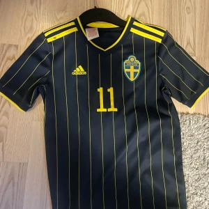 Svensk landslagströja från Adidas - Säljer en snygg svensk landslagströja från Adidas med nummer 11 och namn på ryggen. Tröjan är mörkblå med gula ränder och detaljer, inklusive det svenska landslagets emblem på bröstet. Perfekt för fotbollsfans! 💙💛