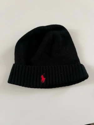 Svart mössa från Polo Ralph Lauren - Svart ribbstickad mössa från Polo Ralph Lauren med klassisk röd logga broderad framtill. Mössan är mjuk och har en bred uppvikt kant som ger en snygg och enkel look. Perfekt för kalla dagar och passar till allt.