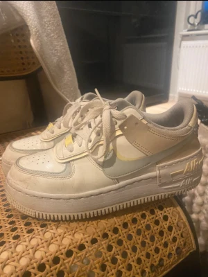Nike Air Force 1 Shadow sneakers - Nike Air Force 1 Shadow sneakers i en ljus färgpalett med inslag av beige, vitt och grått. Skorna har dubbla detaljer, snörning och en chunky plattformssula. Materialet är skinn och syntet, med broderad Nike-logga på hälen och AIR-text på sidan.