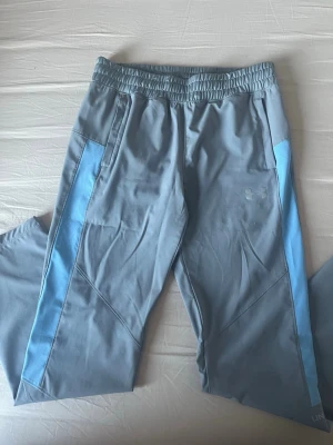 Ljusblå träningsbyxor från Under Armour - Snygga ljusblå träningsbyxor från Under Armour med elastisk midja och dragkedjeförsedda fickor. Byxorna har en cool ljusblå panel längs sidorna och logga framtill. Perfekta för sport eller chill, tillverkade i mjuk polyester som andas.