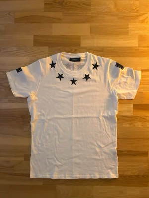 Vit Givenchy vintage t-shirt med stjärnor - Säljer en clean vit t-shirt från Givenchy med svarta stjärnor runt halsen och stora siffror 4 och 7 på ärmarna. Klassisk rund hals och korta ärmar. Tillverkad i mjuk bomull som känns skön mot huden. Perfekt för dig som gillar streetwear med lyxig touch.