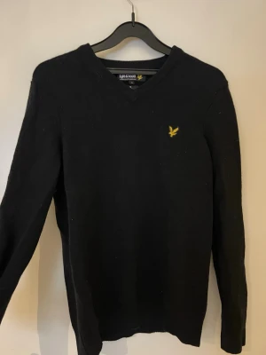 Svart stickad tröja från Lyle & Scott - Svart stickad tröja från Lyle & Scott med v-ringad hals och deras klassiska gula logotyp på bröstet. Tröjan har långärmad design och är gjord i mjukt material som passar perfekt för kyligare dagar. Enkel och stilren modell som funkar till många olika looks.