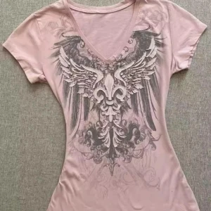 Ljusrosa t-shirt med v-ringning och vingtryck - Ljusrosa t-shirt med v-ringad hals och stort tryck av vingar och ornament på framsidan. T-shirten har korta ärmar och en figurnära passform. Perfekt för dig som vill sticka ut med en cool design.