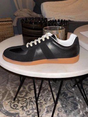 Helt nya Maison Margiela skor - Säljer ett par stilrena helt nya svarta Maison Margiela skor med vit insida och kontrasterande ljusa snören. Skorna har en rund tå och platt gummisula i brun färg. Ovandelen är i en mix av skinn och mocka vilket ger en snygg textur. Perfekt för dig som gillar klassisk och clean stil. HELT OANVÄNDA!