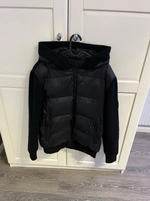 Svart pufferjacka från Evalo med huva - Svart pufferjacka från Bvaldi med snygg huva och stickade ärmar. Jackan har dragkedja framtill och två fickor med dragkedja. Kroppen är quiltad och glansig medan ärmarna är stickade för extra komfort. Perfekt för dig som gillar en modern och sportig look. Storleken är M men sitter som S.