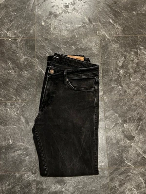 Svarta jeans från Jack & Jones - Säljer ett par svarta jeans från Jack & Jones med klassisk femficksdesign och snyggt läderpatch bak. Jeansen har rak passform och normal midja, tillverkade i slitstarkt bomullsmaterial. Perfekt för dig som gillar stilrena och enkla jeans.