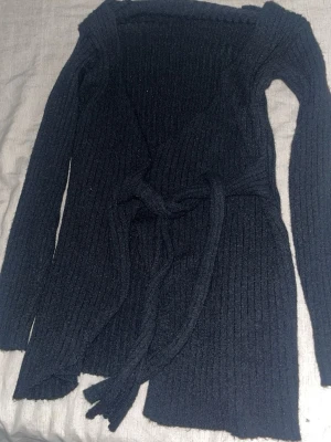 Svart ribbad kofta med knytband - Säljer en svart, långärmad ribbad kofta med knytband i midjan. Koftan har en omlott-design och är gjord i ett stickat material som känns mjukt och skönt. Perfekt att slänga på sig när det är lite kyligt ute.