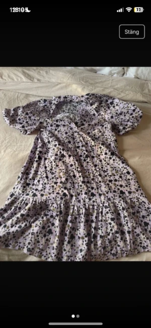 Ljuslila blommig kortklänning C XS - Supersöt ljuslila kortklänning från C i storlek XS med blommigt mönster i svart, vitt och olivgrönt. Klänningen har puffiga korta ärmar, volang nedtill och knappar framtill. Perfekt för dig som gillar romantisk och lekfull stil.