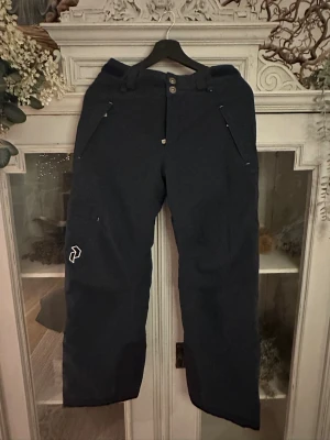 Peak Performance svarta skidbyxor junior - Peak Performance täckbyxor i svart för junior, modell JR THUND P. Byxorna har raka ben, två framfickor med dragkedja och bälteshällor. Materialet är 77% polyester och 23% polyamid, vilket gör dem slitstarka och perfekta för vintern. Snygg logga på benet och bakficka med dragkedja.