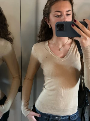 Beige v-ringad tröja Tommy Hilfiger - Snygg beige v-ringad tröja från Tommy Hilfiger med liten logga på bröstet. Tröjan är långärmad, har en slim passform och är tillverkad i mjuk bomullsmix. Perfekt att styla med jeans eller kjol för en clean look.