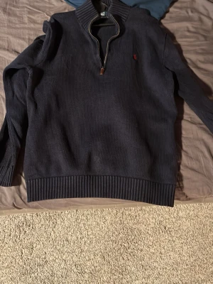 Mörkblå stickad tröja Polo Ralph Lauren - Säljer en mörkblå stickad tröja från Polo Ralph Lauren i storlek M. Tröjan har en half zip-dragkedja vid halsen, ribbade muddar och en röd broderad logga på bröstet. Perfekt för dig som gillar klassisk och stilren design.