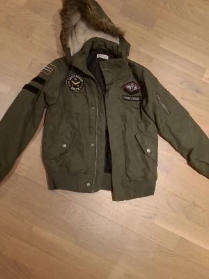 Grön bomberjacka med patchar från H&M - Snygg grön bomberjacka från H&M med coola patchar och emblem på bröstet och ärmen. Jackan har huva med fuskpäls, quiltat svart foder och dragkedja framtill. Två fickor med knappar och en extra ficka med dragkedja på ärmen. Perfekt för dig som gillar streetstyle.