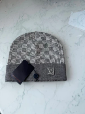 Grå rutig Louis Vuitton mössa - Snygg grå mössa från Louis Vuitton med klassiskt rutigt mönster och uppvikt kant. Mössan har en diskret LV-logga framtill och är stickad i mjukt material som känns lyxigt. Perfekt för dig som gillar exklusiva accessoarer med stilren design.