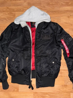 Svart bomberjacka Alpha Industries - Svart bomberjacka från Alpha Industries med grå huva och röd insida. Jackan har dragkedja, ribbade muddar och en snygg patch på bröstet. Klassisk röd tag på ärmen och två fickor framtill. Perfekt för dig som vill ha streetstyle.