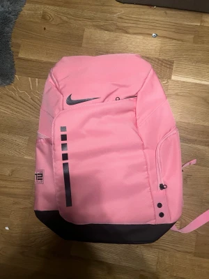 Nike Elite väska  - Säljer en snygg Nike Elite ryggsäck i rosa med svarta detaljer och logga. Väskan har flera fack med dragkedja, justerbara axelremmar och praktisk design. Materialet är slitstark polyester och insidan har meshfickor för smart förvaring. Perfekt för dig som vill sticka ut med färg!