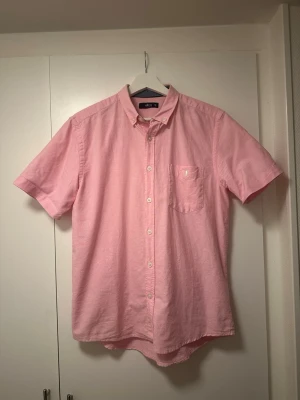 Rosa kortärmad skjorta från Ellos - Snygg rosa skjorta från Ellos i storlek S. Skjortan har korta ärmar, klassisk krage med knappar och en bröstficka. Tillverkad i mjuk bomull som känns skön mot huden. Perfekt för dig som gillar stilrena och färgglada plagg.