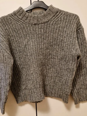(Barnkläder) Grå stickad tröja från Lager 157 - Säljer en grå stickad tröja från Lager 157 med grov ribbad struktur och hög krage. I storlek 140 cm. Tröjan är långärmad och har en loose passform, perfekt för kyliga dagar. Materialet är en mix av akryl, polyester, polyamid och ull som gör den både mjuk och varm.